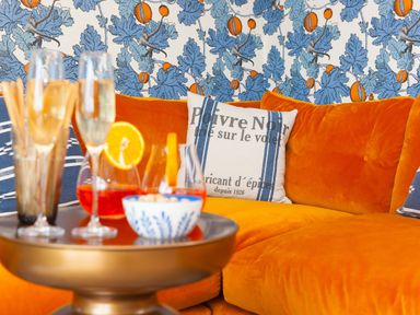 A close-up of an orange velvet sofa with pillows and a small round table with drinks and snacks in a room with blue and orange patterned wallpaper at Sea Sands in Praa Sands