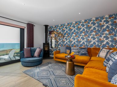 A living room with orange and blue sofas a patterned blue rug a small round table and a window overlooking the sea at Sea Sands Praa Sands