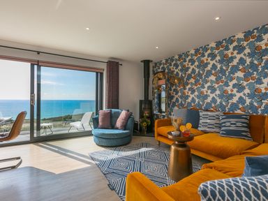A living room with mustard sofas and blue patterned wallpaper overlooking the sea at Sea Sands in Praa Sands