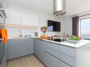 A kitchen with gray lower cabinets white upper cabinets a stovetop on the island and a television on the wall at Sea Sands in Praa Sands