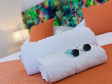 White towels with toiletry bottles on an orange bedspread with orange pillows and a colorful headboard at Sea Sands in Praa Sands