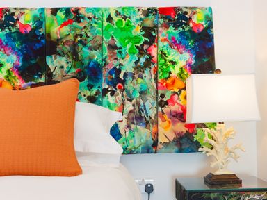 A bed with an orange pillow and a colorful abstract headboard next to a table lamp on a bedside table at Sea Sands in Praa Sands