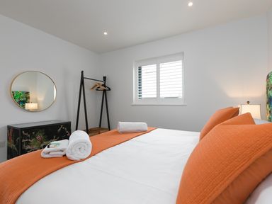 A bedroom with a bed with orange pillows and blanket white towels a black clothes rack with hangers a round mirror and a window at Sea Sands in Praa Sands