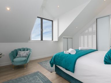A bedroom with a bed with teal bedding and rolled towels a teal armchair near a window with white shutters at Sea Sands in Praa Sands