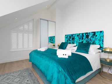 A bedroom with a teal bedspread and matching pillows a patterned headboard a wardrobe with sliding doors a window with shutters and a side table with a lamp at Sea Sands in Praa Sands