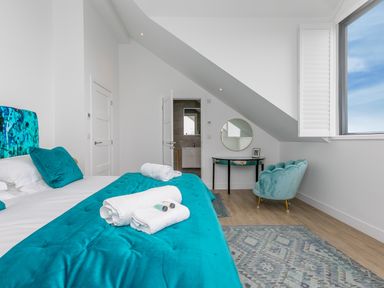 A bedroom with a blue bedspread pillows towels a chair a table with a round mirror and an open window at Sea Sands in Praa Sands