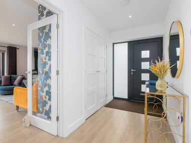 An entrance hall with a black front door a gold-framed mirror and a glass console table next to a doorway showing a living room at Sea Sands in Praa Sands