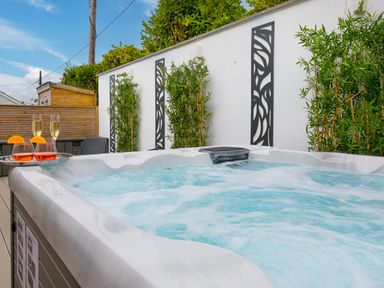 An outdoor hot tub with bubbling water on a wooden deck next to a white wall with decorative panels and plants with drinks on the edge of the hot tub at Sea Sands in Praa Sands