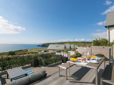 An outdoor patio with a dining table and chairs set with breakfast overlooking the sea and distant hills at Sea Sands in Praa Sands