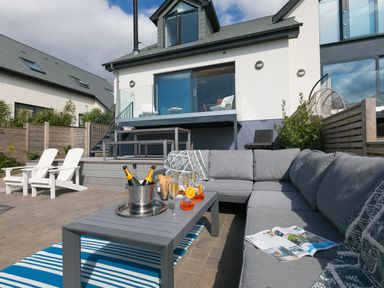 An outdoor patio with gray sectional sofa and table with drinks and bottles in front of a house with balcony at Sea Sands in Praa Sands