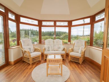 A seating area with wicker furniture and cushions by large windows overlooking greenery at Redford View in Culdaff County Donegal