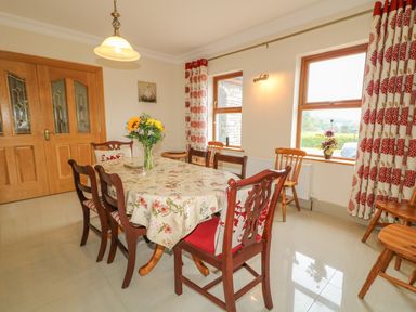 A dining room with a table covered with a floral tablecloth and wooden chairs near windows with red patterned curtains at Redford View in Culdaff County Donegal