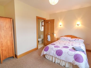 A bedroom with a double bed covered with a duvet with purple circles a wooden wardrobe a door leading to a bathroom and beige walls at Redford View in Culdaff County Donegal