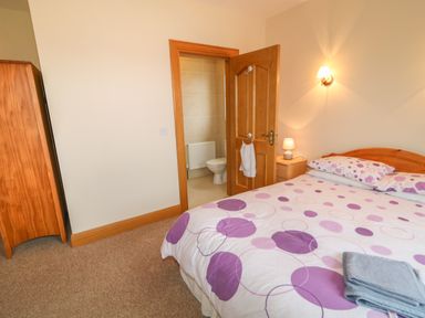 A bedroom with a bed with purple polka dot bedding a wooden wardrobe a nightstand with a lamp and an open door leading to a tiled bathroom at Redford View in Culdaff County Donegal