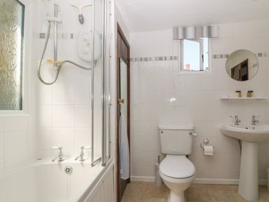 A bathroom with a bathtub and shower on the left a toilet in the middle and a sink with a round mirror above it on the right at Pixie Cottage in Looe