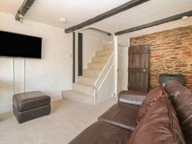 A living room with a leather sofa a ottoman a wall mounted television and stairs at Pixie Cottage in Looe