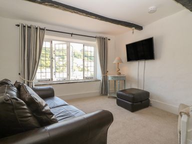 A living room with a leather sofa a window with curtains a side table with a lamp a wall-mounted TV and an ottoman at Pixie Cottage in Looe