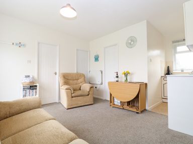 A living room with beige armchairs a wooden folding table with flowers and wine near a kitchen at 14 Siesta Mar in Mundesley