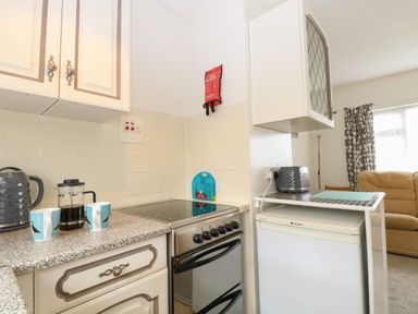 A kitchen area with a stove, fridge, kettle, coffee pot, two mugs, and a toaster with a living room visible in the background at 14 Siesta Mar in Mundesley