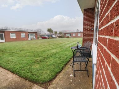 A small outdoor seating area with a table and two chairs next to a brick building overlooking a grassy courtyard with other brick buildings and parked cars at 14 Siesta Mar in Mundesley
