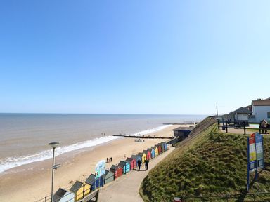 A beach with colorful beach huts along a walkway and a grassy hill with benches by the sea at 14 Siesta Mar in Mundesley