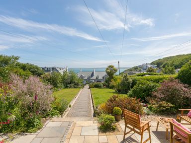 A garden with lawn and flower beds a pathway and wooden chairs overlooking houses and the sea at 6 Albany Terrace in St Ives