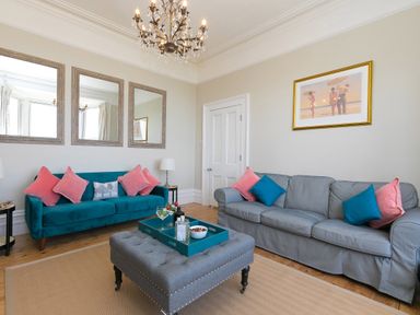 A living room with blue and gray sofas pink and blue cushions a gray ottoman with a tray on it three mirrors on the wall and a framed picture at 6 Albany Terrace in St Ives