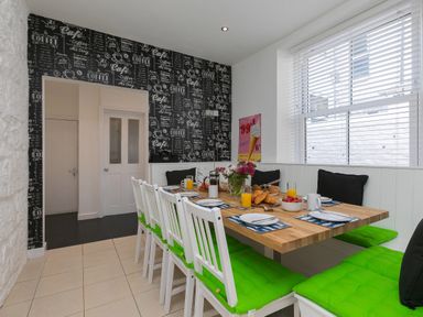 A dining area with a wooden table set for breakfast white chairs with green cushions and a corner bench with black cushions at 6 Albany Terrace in St Ives