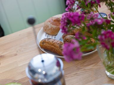 A wooden table with a plate of bread rolls a French press and pink flowers in a glass vase at 6 Albany Terrace in St Ives