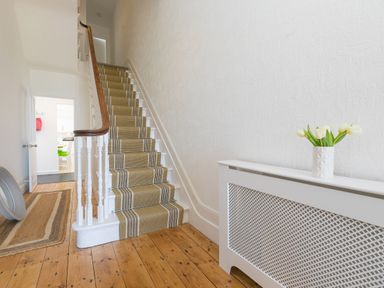 A stairway with a striped runner carpet and white railings with a wooden handrail next to a white radiator cover with a vase of flowers at 6 Albany Terrace in St Ives