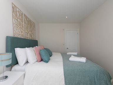 A bedroom with a bed with teal headboard and cushions white bedside table with lamp and white door at 6 Albany Terrace in St Ives