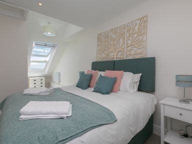 A bedroom with a double bed with pillows and folded towels a bedside table with a lamp and a small chest of drawers near a window at 6 Albany Terrace in St Ives