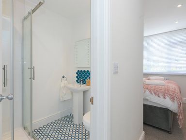 A bathroom with a glass shower door, pedestal sink, white walls, and patterned blue and white floor tiles next to a bedroom with a bed and folded towels at 6 Albany Terrace in St Ives