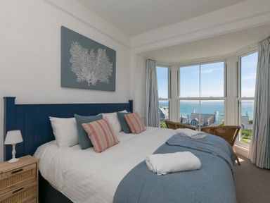 A bedroom with a large bed pillows and towels next to a bay window overlooking houses and the sea at 6 Albany Terrace in St Ives