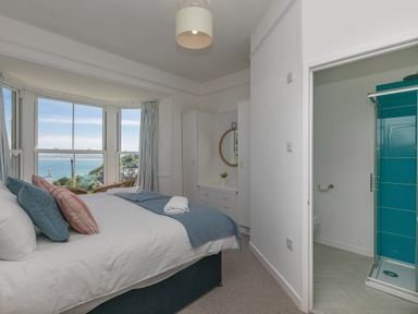 A bedroom with a bed facing a bay window overlooking the sea a dressing table with a round mirror and an ensuite bathroom with a shower at 6 Albany Terrace in St Ives