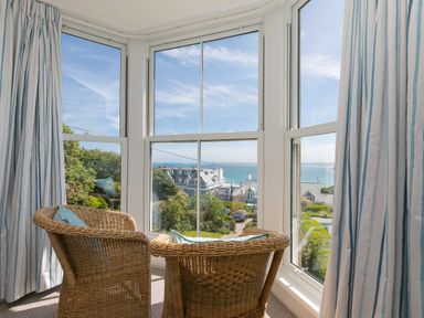 Two wicker chairs with cushions facing large windows overlooking houses trees and ocean at 6 Albany Terrace in St Ives