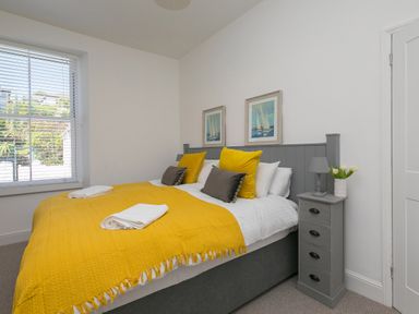 A bedroom with a bed with yellow and gray pillows and a gray bedside cabinet with a lamp and flowers at 6 Albany Terrace in St Ives