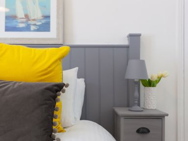 A bed with yellow and gray pillows next to a gray bedside table with a lamp and vase with flowers at 6 Albany Terrace in St Ives