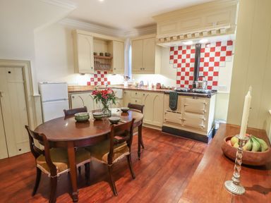 A kitchen with a wooden dining table and chairs beige cabinets and a white stove with red and white tiled backsplash at Lion House in Moretonhampstead