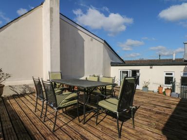 An outdoor wooden deck with a metal table and six chairs with cushions at Lion House in Moretonhampstead