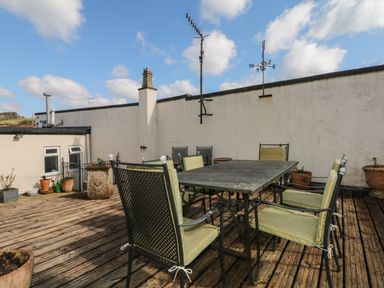 An outdoor patio with a metal table and six metal chairs with cushions and potted plants on wooden decking at Lion House in Moretonhampstead