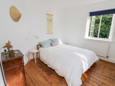 A bedroom with a double bed white bedding wooden floor a wooden chest of drawers and an open window at Tor Cottage in Colwall Herefordshire