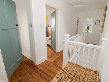 A hallway with wooden floors a wicker bench white walls a white banister and green doors at Tor Cottage in Colwall Herefordshire
