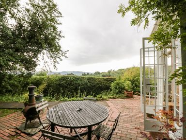 A patio with a round wooden table and chairs near open glass doors and plants at Tor Cottage in Colwall Herefordshire