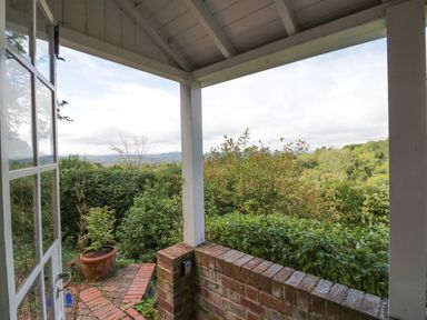 A covered porch with brick railing overlooking a garden with bushes and trees at Tor Cottage in Colwall Herefordshire
