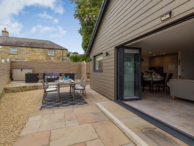 An outdoor patio area with a dining table and chairs next to an open door leading to an indoor dining and living space at Secret Kingdom Cottage in Warkworth