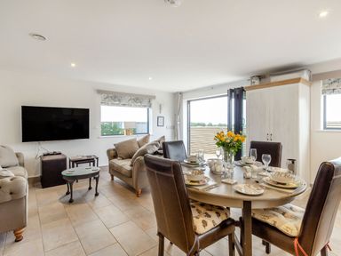 A living and dining area with a round table set for four people a sofa a wall-mounted television and a cupboard at Secret Kingdom Cottage in Warkworth