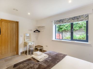 A bedroom with a bed, a wooden door, a small dressing table with a mirror, stools, a window with a floral blind, and a wall shelf with hangers at Secret Kingdom Cottage in Warkworth
