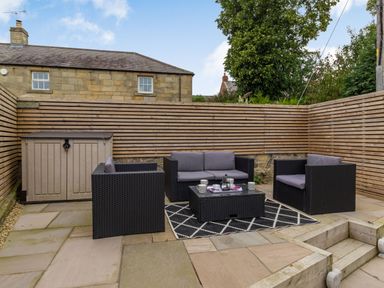 An outdoor patio with black wicker furniture including a sofa, two armchairs, and a coffee table on a patterned rug with a wooden fence and a storage shed in the background at Secret Kingdom Cottage in Warkworth