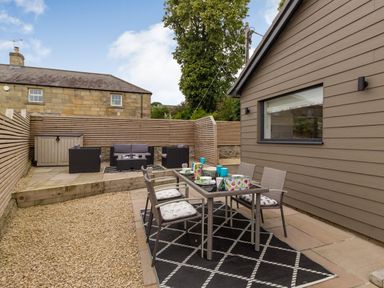 An outdoor patio with a dining table and chairs on a rug and a seating area with sofa and chairs behind a wooden fence at Secret Kingdom Cottage in Warkworth